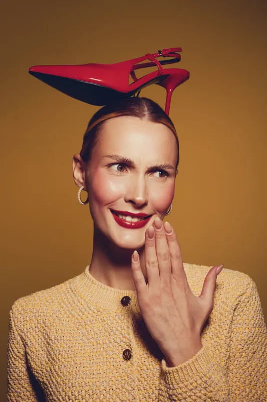 Smiling woman with red high heel shoe balanced on her head wears yellow textured jacket and gold hoop earrings. Studio portrait on brown background with playful and quirky fashion concept.