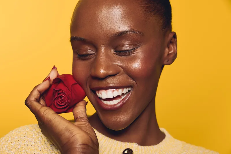 Smiling dark skinned African woman with natural skin holds red rose near face. Yellow background and soft lighting create warm mood. Concept of beauty, joy, and floral elegance.