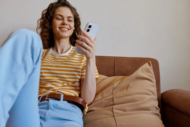 A relaxed woman sits on a couch at home, scrolling her phone and smiling, a casual lifestyle moment. Cozy living room with soft daylight, capturing everyday comfort and connection.