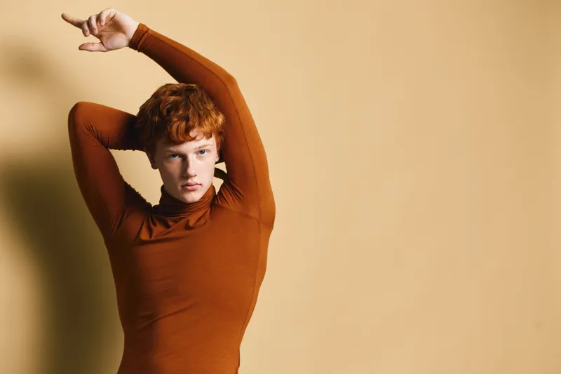 Young person with red hair wearing brown turtleneck sweater posing with arms raised against beige studio background, fashion portrait with soft lighting and serious expression.