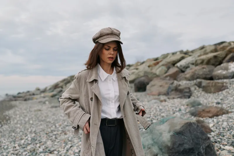Young woman in beige coat and hat walks on rocky beach under cloudy sky. Casual autumn fashion with natural makeup in outdoor coastal environment. Concept of style and solitude.