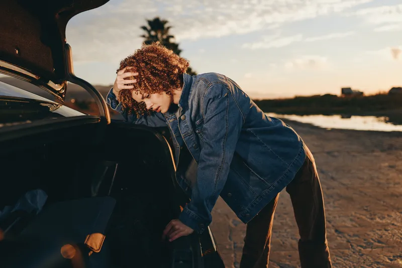 Curly hair woman in denim leaning into a car outdoors at sunset, golden sky behind, adjusting jacket before travel, lifestyle roadtrip moment, candid warm sunlight, evening wind