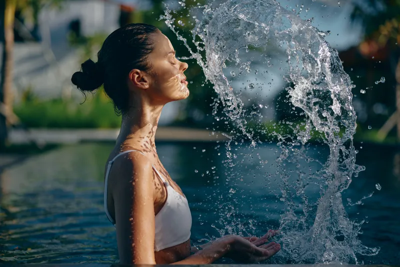 Young woman in white swimsuit enjoying water splash outdoors, eyes closed, sunlit skin, relaxed mood, summer lifestyle, natural background with soft focus.