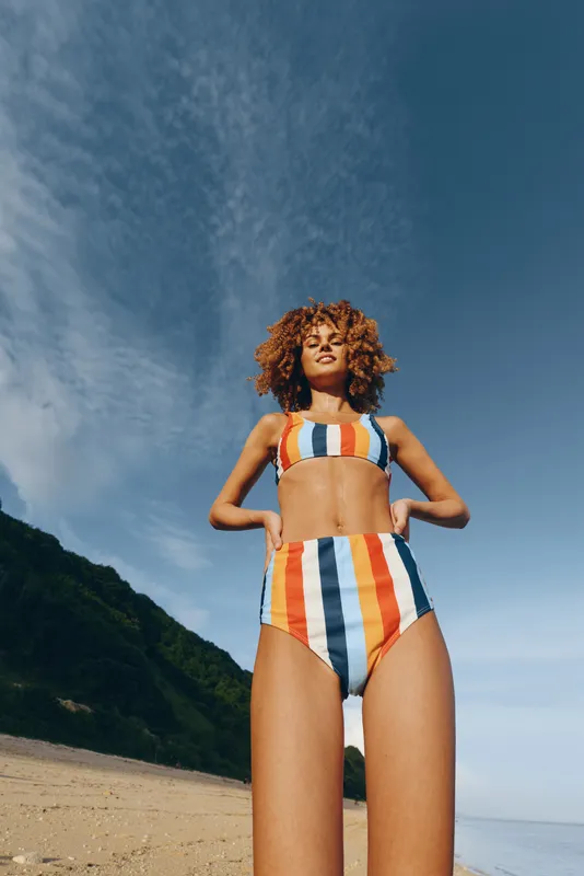 Beach woman in bikini standing confidently on sandy coast under clear blue sky in summer vacation, ocean horizon and green hills behind her body positivity curly hair and relaxed pose