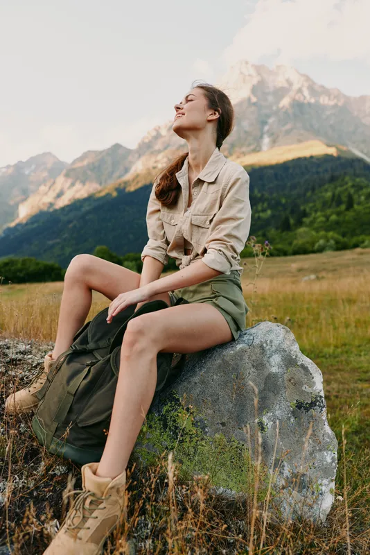 A woman rests on a rock during an outdoor hike, eyes closed, soaking sunlight, with a backpack nearby in a scenic mountain meadow, conveying calm and adventure.