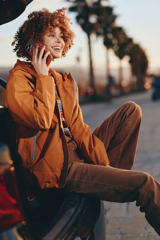 Smiling woman with curly hair, wearing an orange jacket and jeans, sitting on a sidewalk in an urban street, warm daylight, relaxed lifestyle mood, outdoors, soft bokeh background today