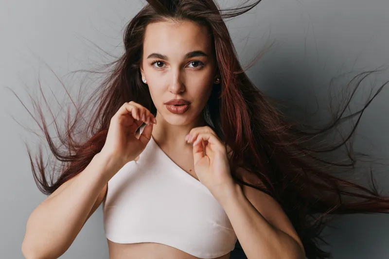 Young woman with long flowing hair wearing white top posing against gray background. Female model with natural makeup and confident expression in studio portrait photography.