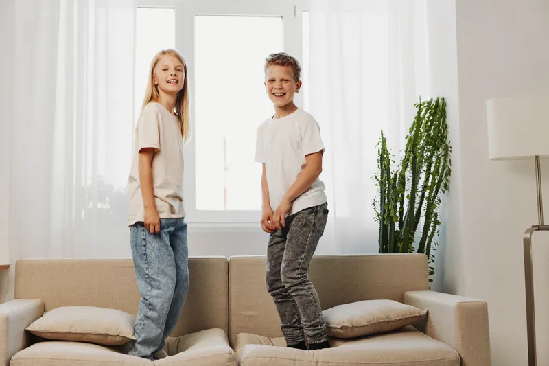 Two happy children in casual clothes jump on a beige sofa in a bright living room with natural light and green plant. Concept of childhood joy and playful moments.