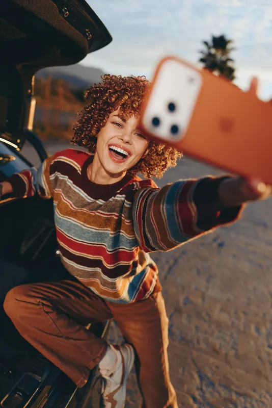 Woman selfie smile curly hair outdoors car lifestyle, young adult cheerful expression, striped sweater, leaning near open vehicle door, warm sunlight, urban summer travel relaxed mood