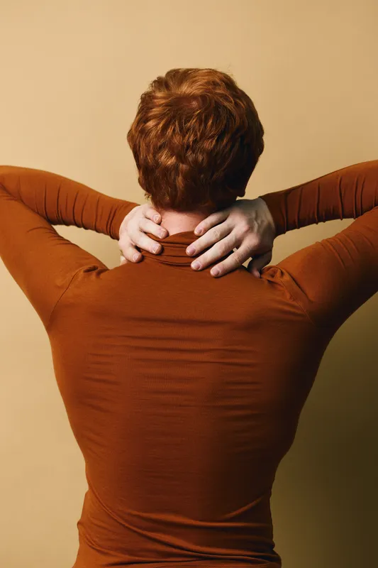 Person with short hair and brown turtleneck sweater stretching neck with hands behind head on neutral background for relaxation and self care concept.