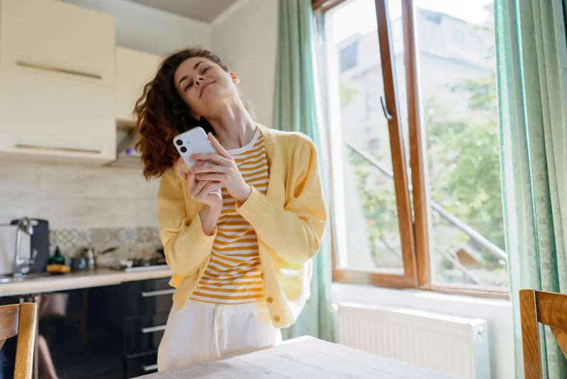 Cheerful woman at home, in a sunlit kitchen, glancing upward with a smile while holding a phone, wearing a striped tee and yellow cardigan, casual lifestyle moment, soft natural light, warm domestic