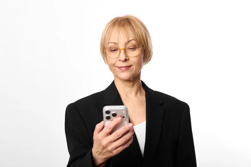 Senior woman with glasses holding smartphone, business blazer on white background, close portrait, calm smile, mobile communication, modern technology concept, soft studio light