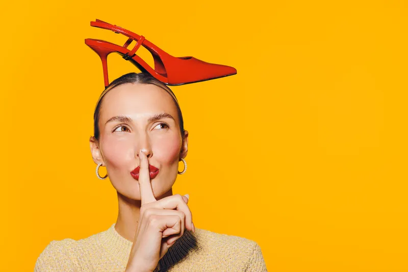 Young woman with red high heel shoe on head holds finger to lips in shushing gesture. Bright yellow background creates playful and quirky fashion concept with confident expression.