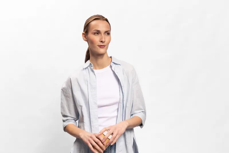 Young woman with light brown hair wearing casual white shirt and grey overshirt stands against white background. She looks calm and thoughtful with hands clasped in front. Concept of simplicity.