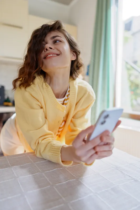 Smiling woman at home leaning over a table, taking a selfie with a smartphone. Cozy kitchen setting, casual outfit, warm natural light, relaxed lifestyle and self expression.