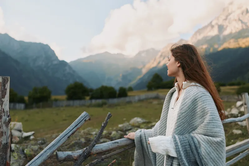 Young woman with long hair gazes at alpine mountains, wrapped in a cozy shawl, standing beside a weathered wooden fence, enjoying a calm lifestyle moment in nature