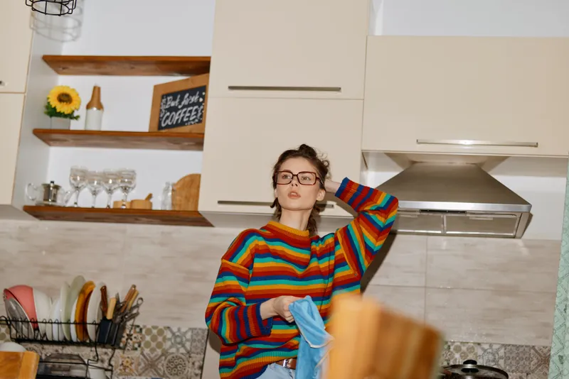 Casual woman in a bright home kitchen drying dishes after cooking, lifestyle portrait, cozy interior, everyday chores, relaxed mood, colorful striped sweater, tidy countertops and shelves.