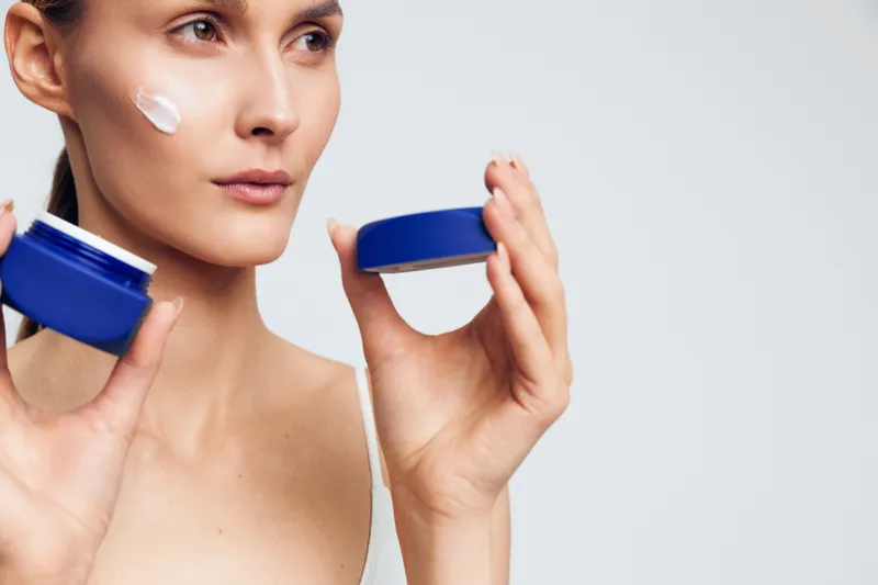 Young woman applying face cream with a clean skin and neutral expression. Close-up on a white background. Concept of skincare, beauty, and daily routine.