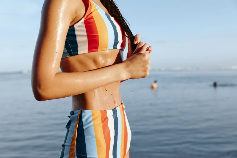 Close up of woman at the beach wearing a striped summer dress adjusting the neckline in bright sunlight, ocean backdrop suggests vacation fashion and outdoors lifestyle, natural warm light