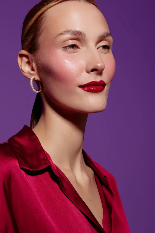 Close-up portrait of a confident woman with red lipstick and shiny skin wearing a red satin blouse and hoop earrings on purple background. Concept of beauty, fashion, and elegance.