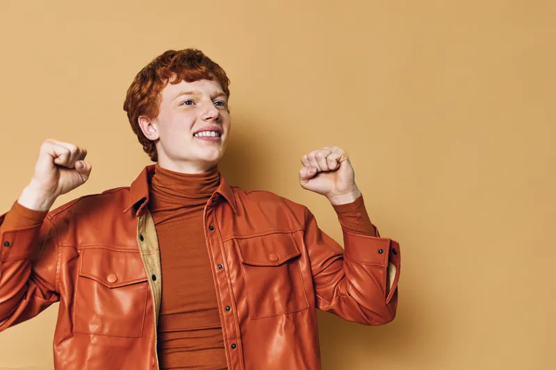Young man with red hair wearing orange leather jacket and turtleneck raises hands in celebration against beige background with happy facial expression and confident posture.
