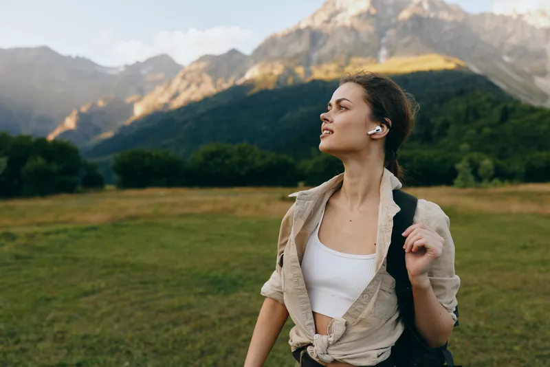 Young woman with short hair wears a white crop top and beige shirt as she hikes a mountain meadow, gazing up with a calm smile under clear blue skies, sunlight highlighting her relaxed posture and