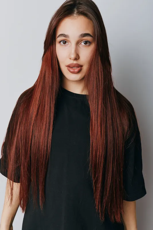 Young woman with long straight red hair and natural makeup wearing black t shirt standing against light background looking at camera with confident expression and relaxed posture.