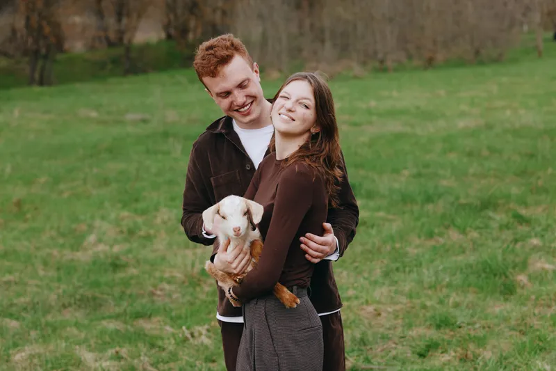 Young couple embraces outdoors while the woman holds a small baby goat in her arms on a green grassy field during a casual lifestyle moment.