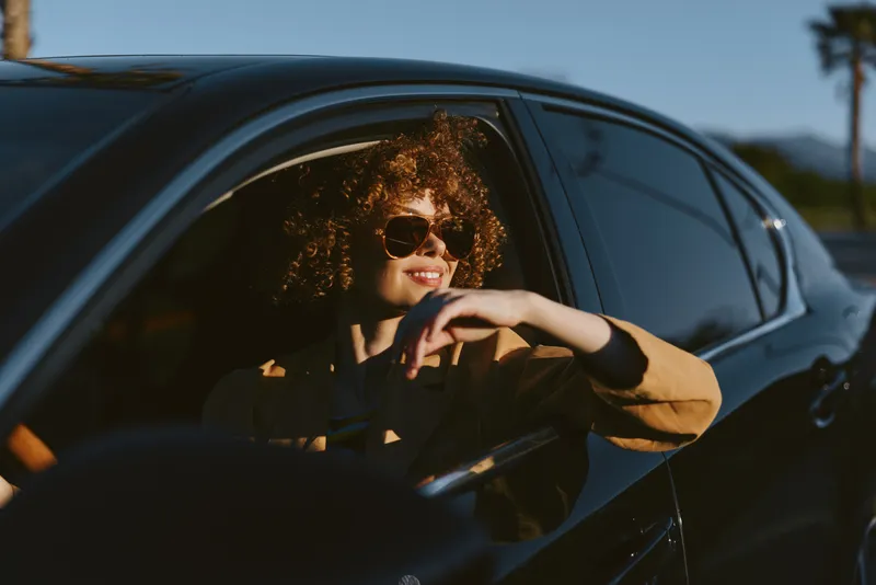 Woman driver sits inside a sleek car with sunglasses, relaxed smile, enjoying a sunny day, casual roadside scene, retro film color mood, lifestyle portrait through the window
