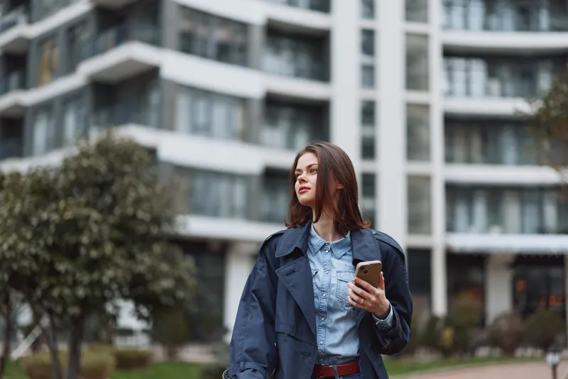 Young woman in denim jacket stands outdoors near modern apartments, holding a smartphone, thoughtful expression, casual urban portrait in daytime light, soft weather, relaxed mood, sunny.