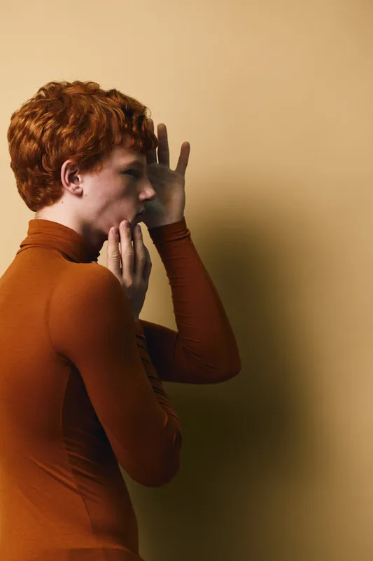 Woman with short curly hair and brown turtleneck covering mouth by hand whispering to wall with dramatic lighting and neutral background for secret or message concept.