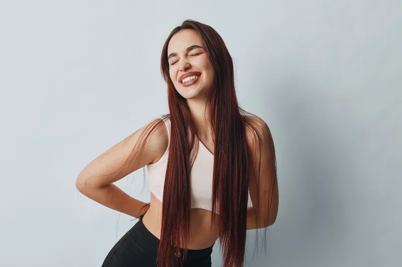 Young woman with long hair in sportswear smiling with eyes closed and hands on back for pain relief or stretching against light background in studio.
