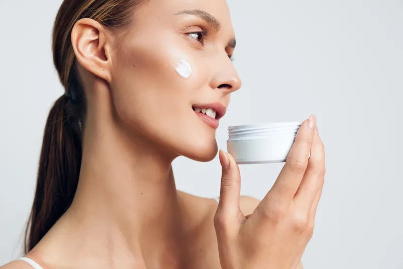A woman with brunette hair applies moisturizer from a white jar, close up in a clean studio, capturing a skincare moment of self care and calm.