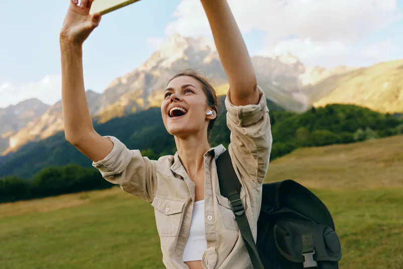 Joyful woman outdoors, raising a phone for a selfie, backpacked traveler, mountain backdrop, sunny day, carefree lifestyle, natural colors, expansive landscape