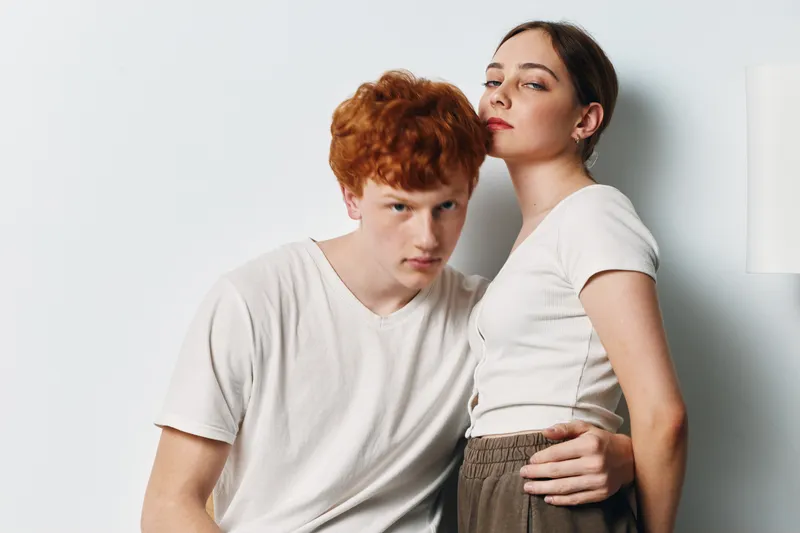 Young man and woman in casual clothing posing together with confident expressions in studio. Redhead male and brunette female standing close in white t shirts on white background.