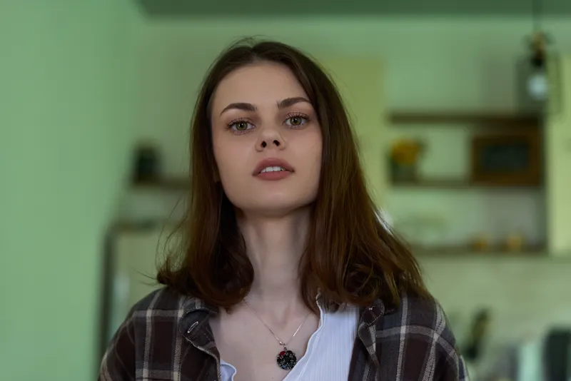 A woman in a casual plaid shirt stands indoors, facing the camera with a calm, thoughtful expression. A cozy home setting hints at everyday lifestyle and confidence