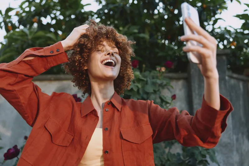 Cheerful woman outdoors, selfie moment, curly hair, jacket, garden background, sunlight glow, casual lifestyle, natural colors, relaxed pose, candid joy