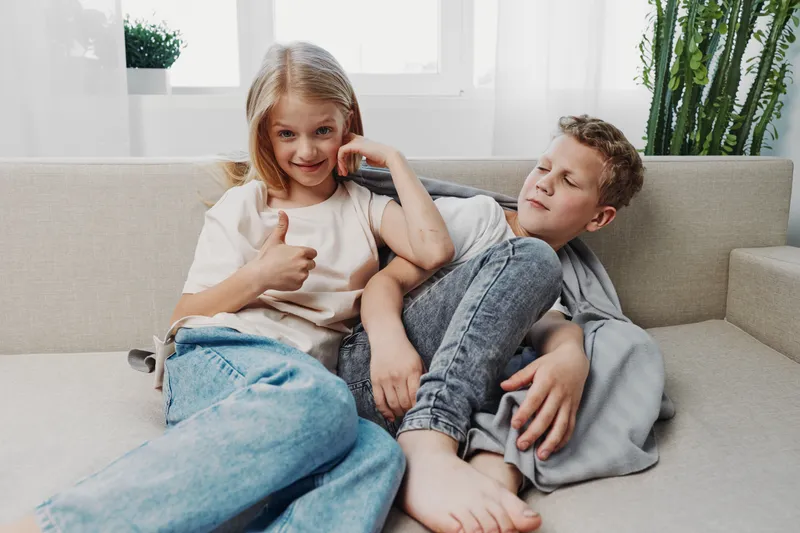 Happy girl giving thumbs up while sitting on couch next to sleepy boy resting with eyes closed. Casual home interior with natural light shows sibling bond and relaxed family moment.