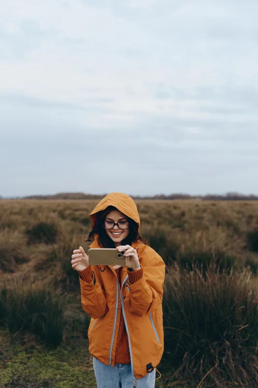 Woman lifestyle outdoors autumn field, smiling, orange jacket with hood, rural countryside landscape, holding camera device, casual portrait, daytime scenic background, natural soft light