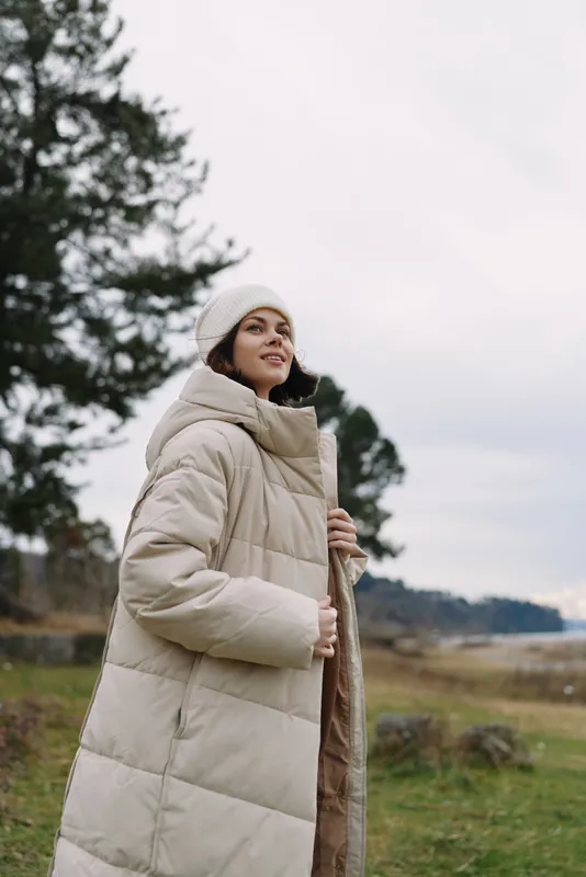 Outdoor woman in a beige winter coat and beanie stands in a tranquil countryside landscape, embracing cold weather and calm nature