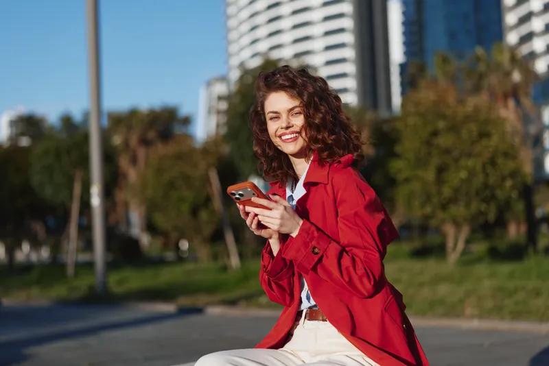 Young woman with curly hair in a red jacket sits outdoors in an urban park, smiling while checking her phone, capturing a bright candid lifestyle moment.
