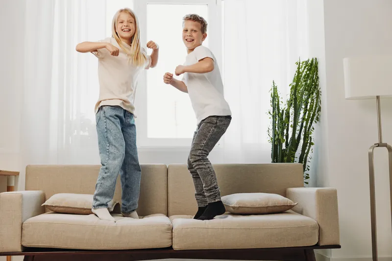 Happy children jumping on sofa in living room, smiling and playing actively. Bright interior with plants and cushions creates joyful home atmosphere.
