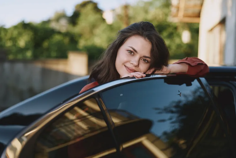 Smiling woman leaning on a car window outdoors, casual lifestyle moment, sunshine on glass, natural candid portrait conveying happiness and relaxed travel mood