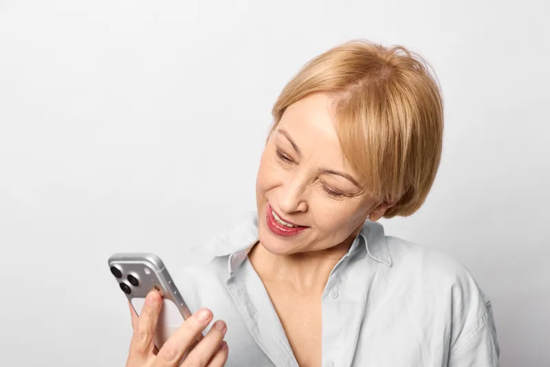 Elderly woman smartphone communication smile digital messaging portrait home technology, she holds a mobile screen, relaxed smile while reading messages, closeup indoors on clean white background