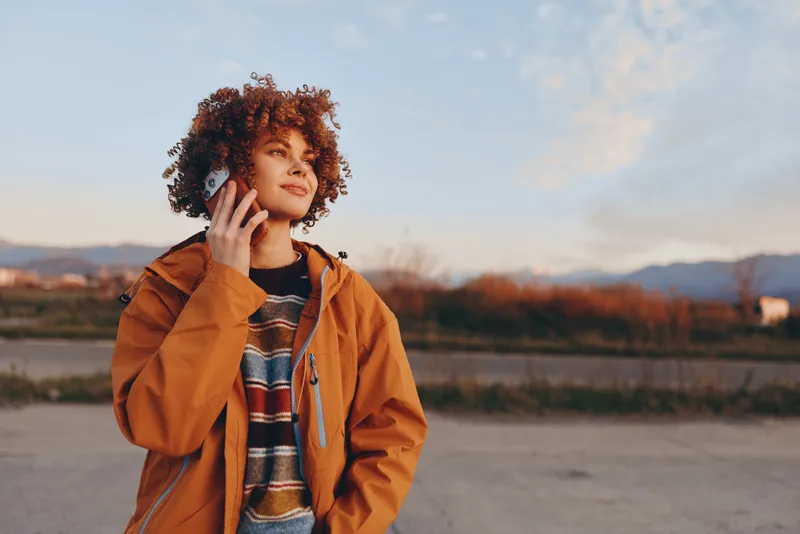 Woman outdoor lifestyle, curly hair warm smile, orange jacket and striped sweater, hand near face, soft breeze, autumn evening by river, distant mountains, natural light