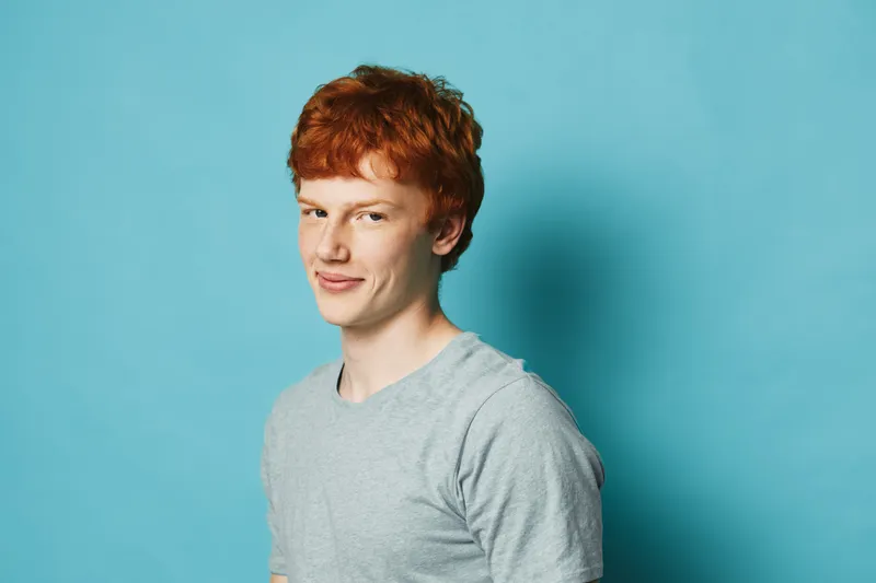 Red hair young man with light skin wearing gray t shirt posing smiling against blue background looking at camera with confident expression and relaxed posture.