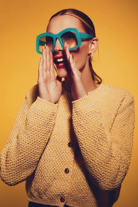 Young woman in yellow sweater and large blue sunglasses shouts or calls out with hands near mouth. Bright studio lighting and orange background create energetic, playful mood.