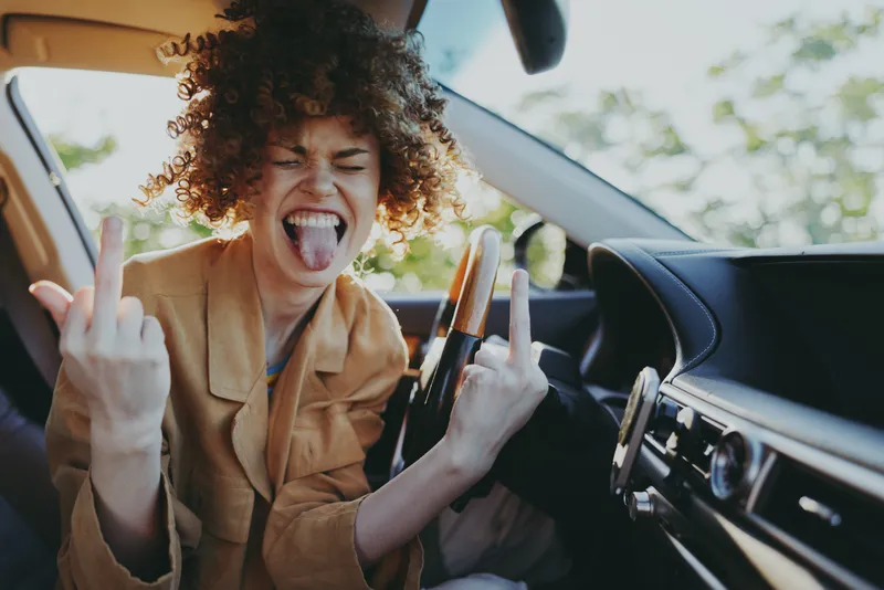 Vibrant woman in a car sticks out her tongue and makes a bold gesture, capturing rebellious street style and carefree attitude inside a sunlit cabin, with dramatic shadows and bright highlights.