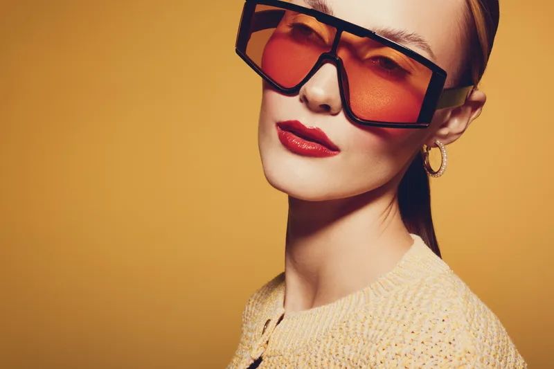 Stylish woman with red lipstick and oversized orange sunglasses poses against yellow background. Fashion portrait with trendy accessories and confident expression. Concept of modern style and beauty.