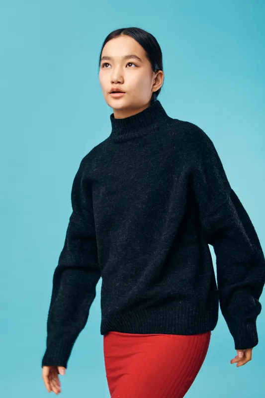 Young woman wearing black turtleneck sweater and red skirt poses against blue background. Casual fashion, female model with natural makeup and simple hairstyle looking sideways in studio.